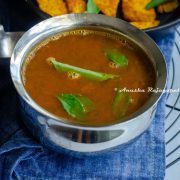 tomato rasam served in a steel bowl placed over a blue napkin. Fried yam slices at the background