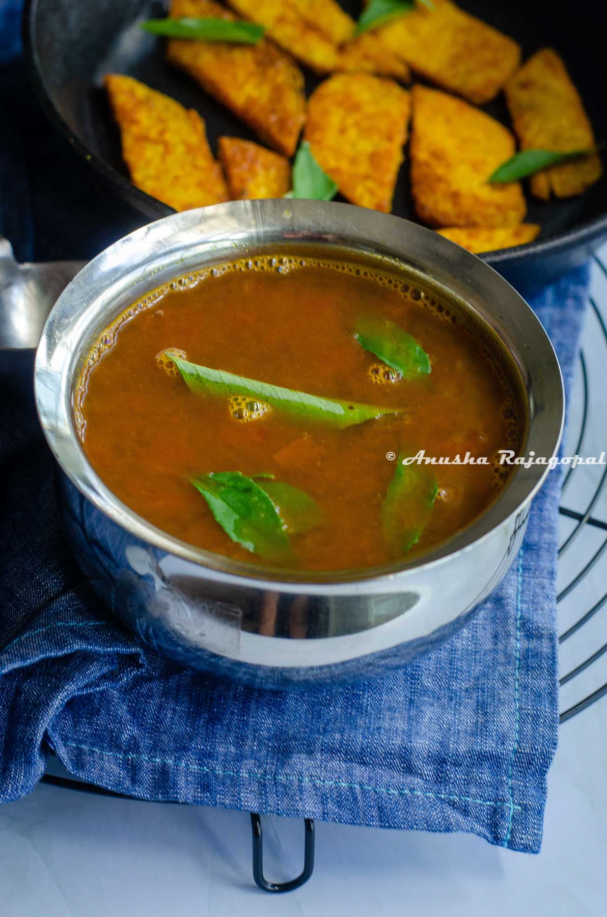 tomato rasam served in a steel bowl placed over a blue napkin. Fried yam slices at the background