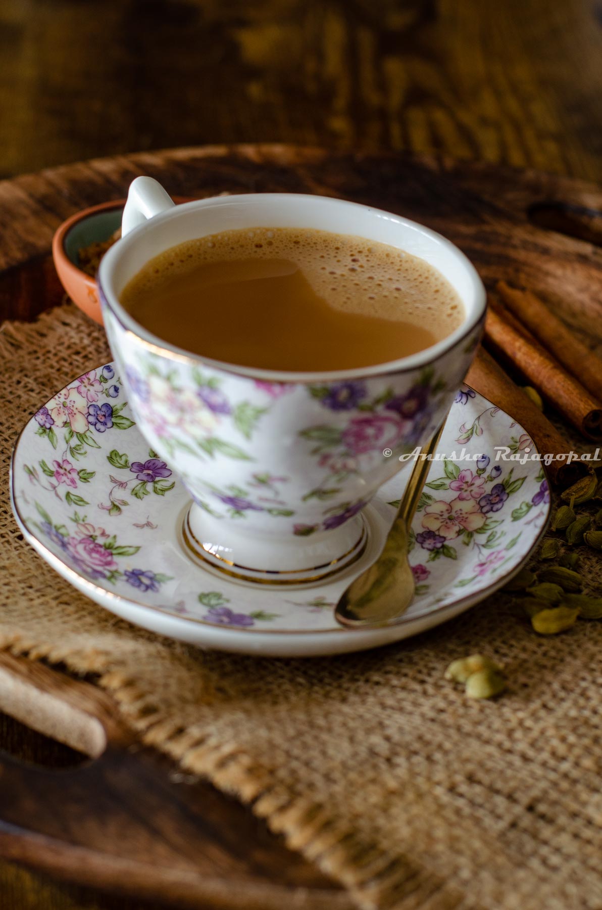 vegan masala chai served in a floral patterned cup and saucer set. A small sugar spoon rests on the saucer. A few cinnamon sticks, a pinch bowl of sugar and some whole spices by the side and around the cup of tea.