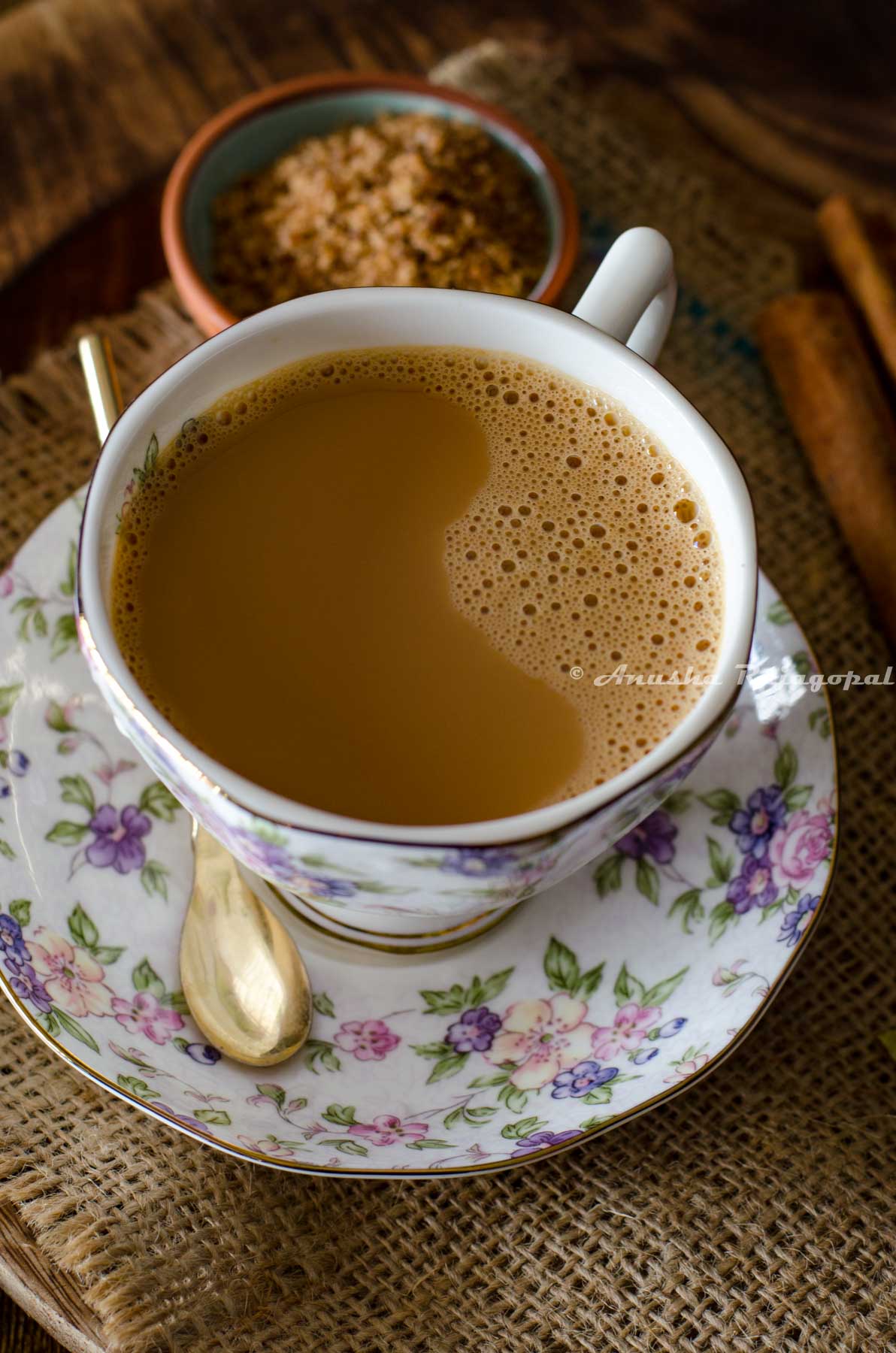 vegan masala chai served in a floral patterned cup and saucer set. A small sugar spoon rests on the saucer. A few cinnamon sticks, a pinch bowl of sugar and some whole spices by the side and around the cup of tea.