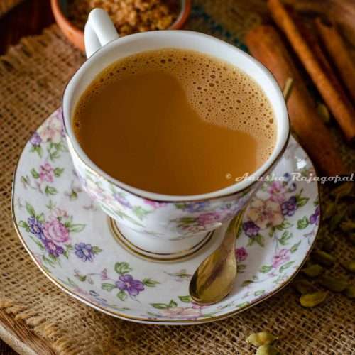 vegan masala chai served in a floral patterned cup and saucer set. A small sugar spoon rests on the saucer. A few cinnamon sticks, a pinch bowl of sugar and some whole spices by the side and around the cup of tea.