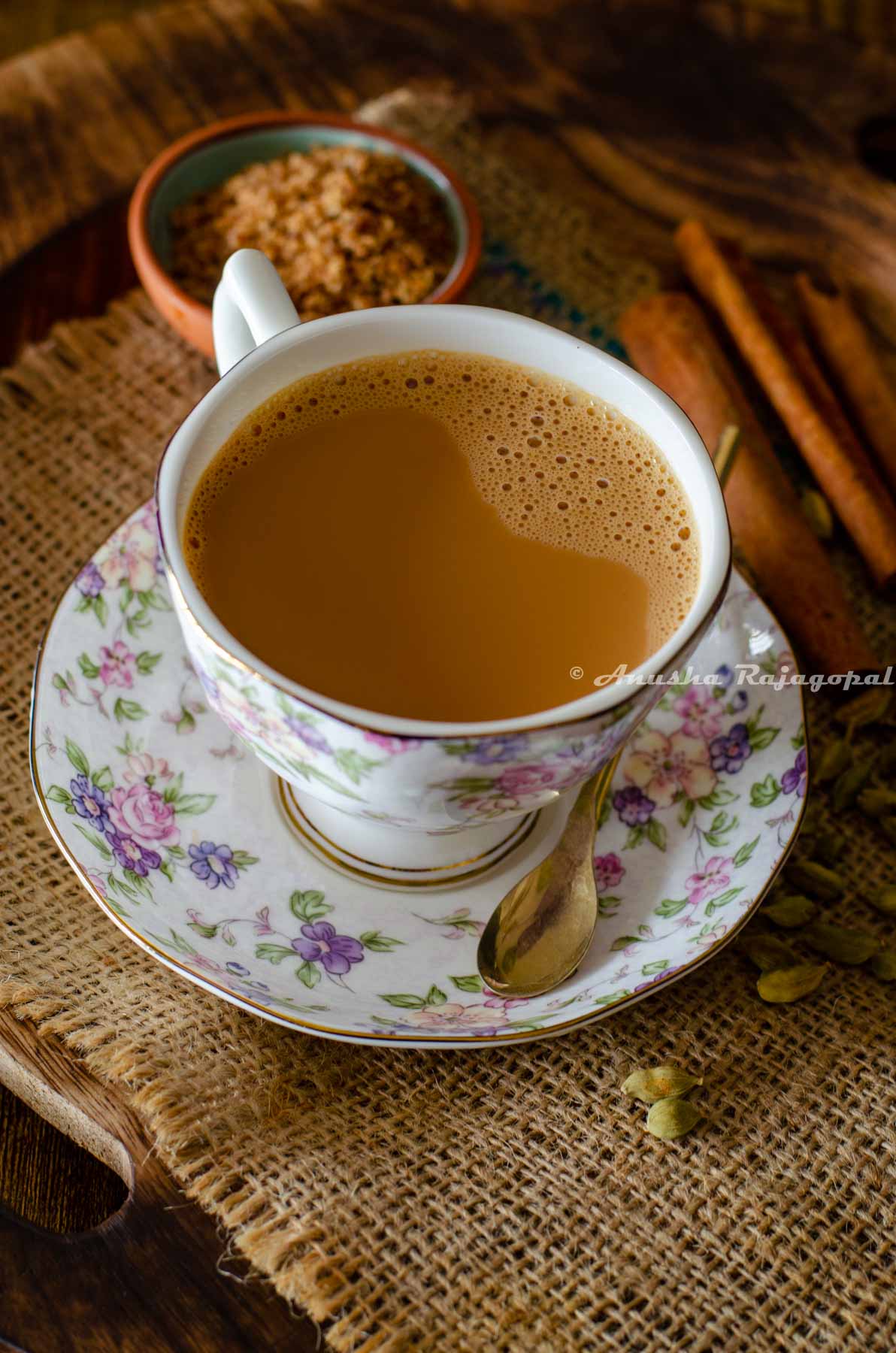 vegan masala chai served in a floral patterned cup and saucer set. A small sugar spoon rests on the saucer. A few cinnamon sticks, a pinch bowl of sugar and some whole spices by the side and around the cup of tea.