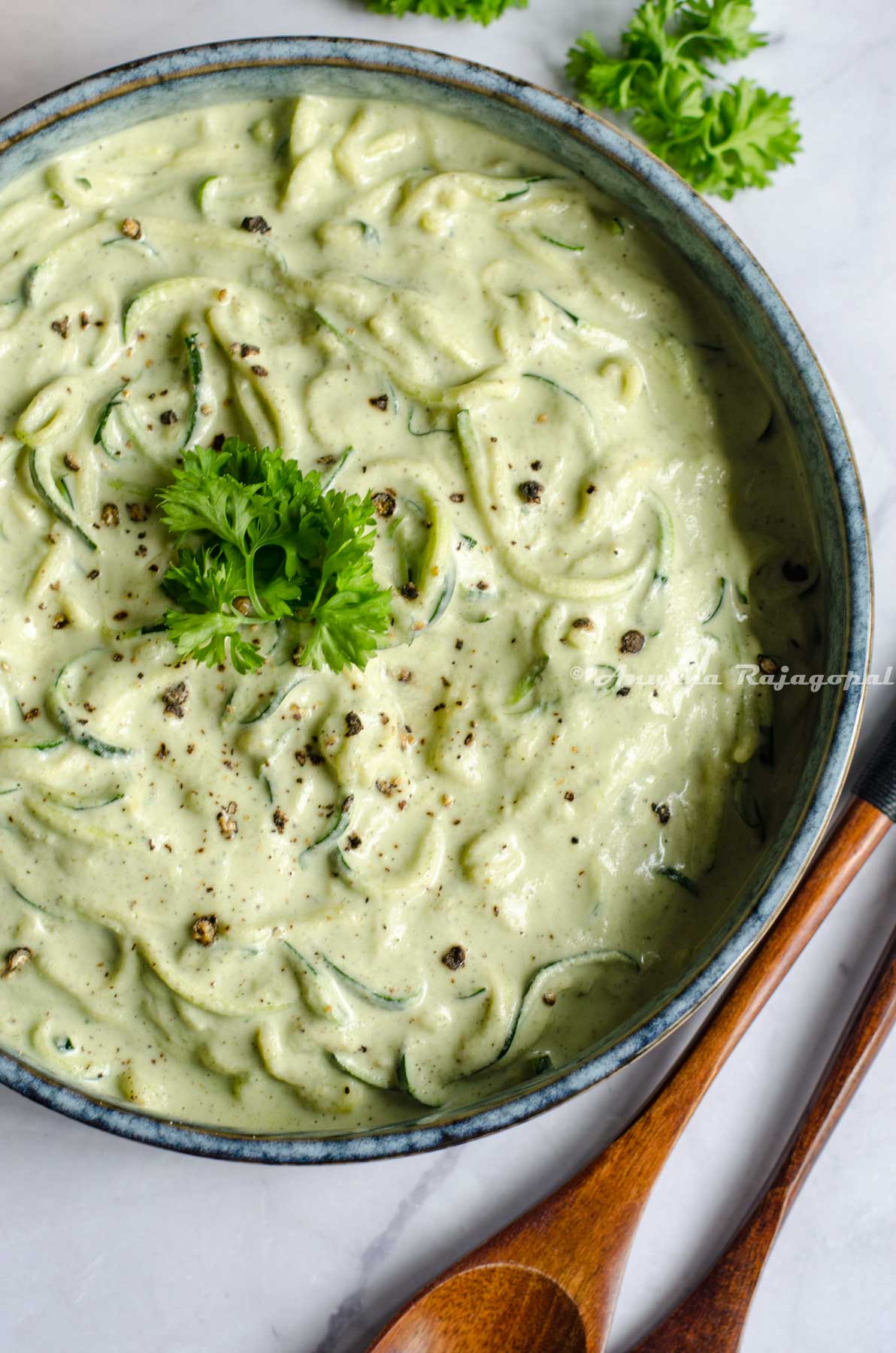 vegan zucchini spaghetti in a greyish rimmed shallow bowl placed on a white marble table top. Spoons, herbs and lemon by the side of the bowl.