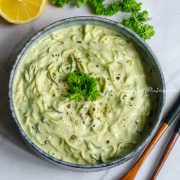 vegan zucchini spaghetti in a greyish rimmed shallow bowl placed on a white marble table top. Spoons, herbs and lemon by the side of the bowl.