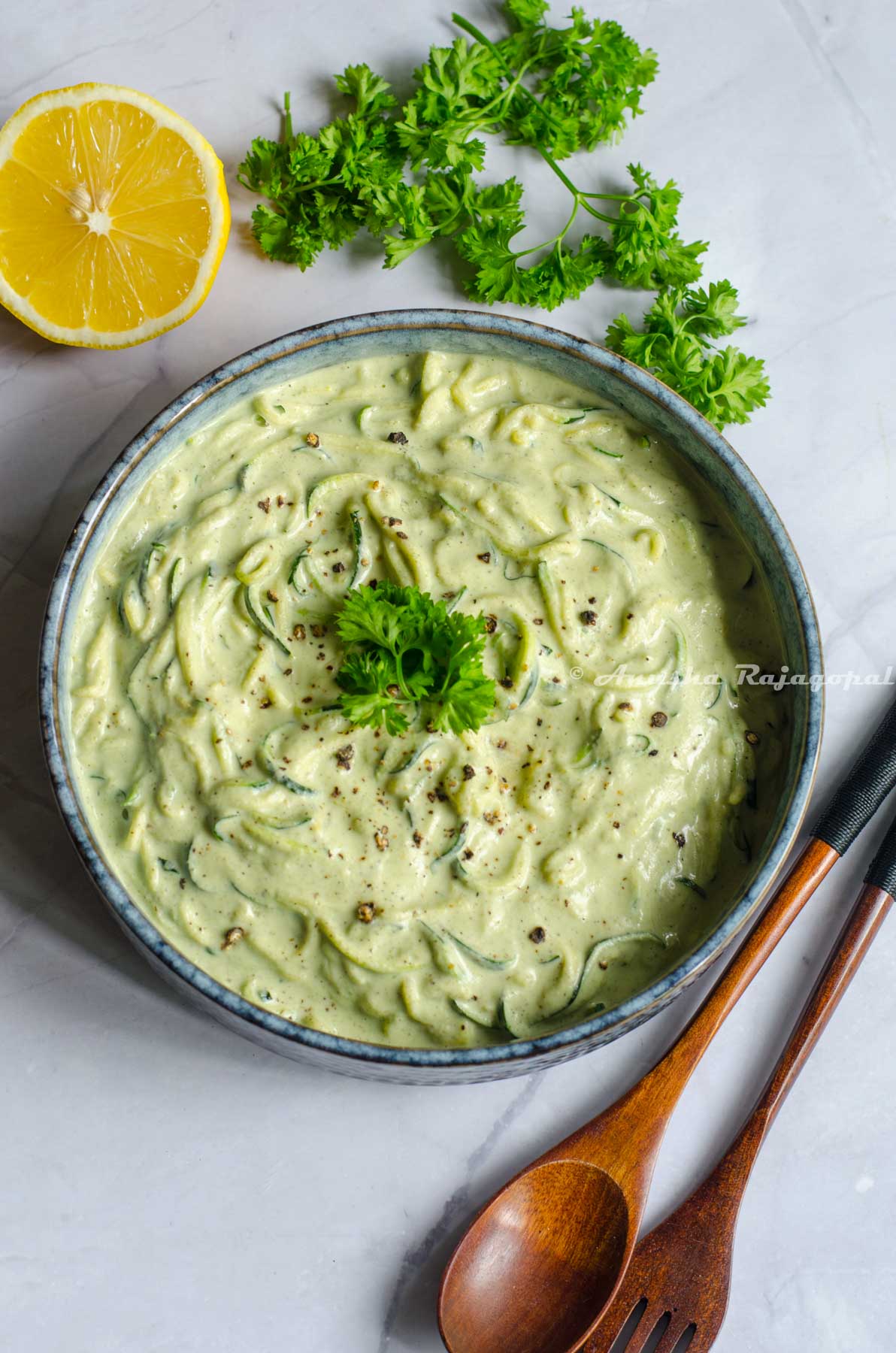 vegan zucchini spaghetti in a greyish rimmed shallow bowl placed on a white marble table top. Spoons, herbs and lemon by the side of the bowl.