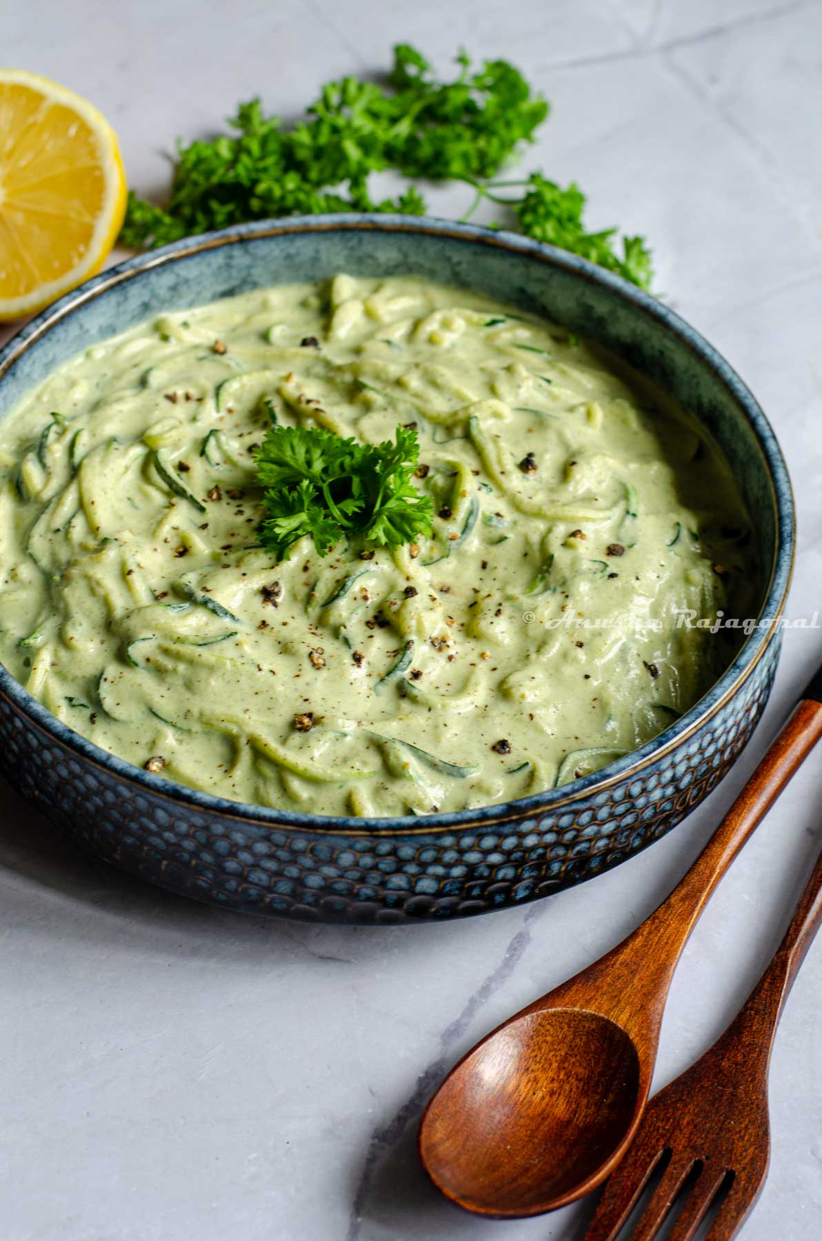 vegan zucchini spaghetti in a greyish rimmed shallow bowl placed on a white marble table top. Spoons, herbs and lemon by the side of the bowl.