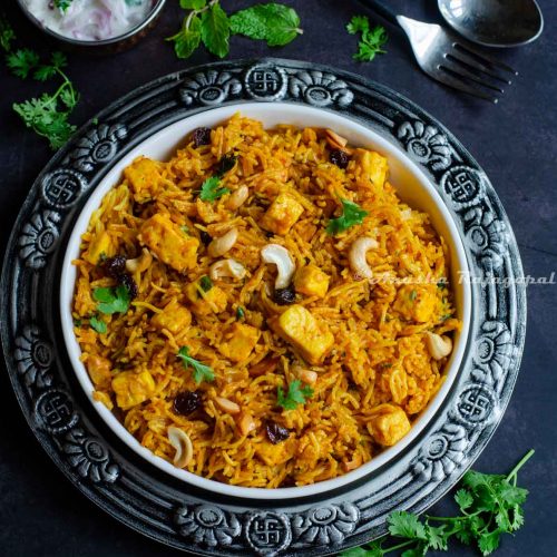 a-bowl-of-paneer-biryani-served-in-a-white-platter-with-onion-raita-and-cutlery-by-the-side