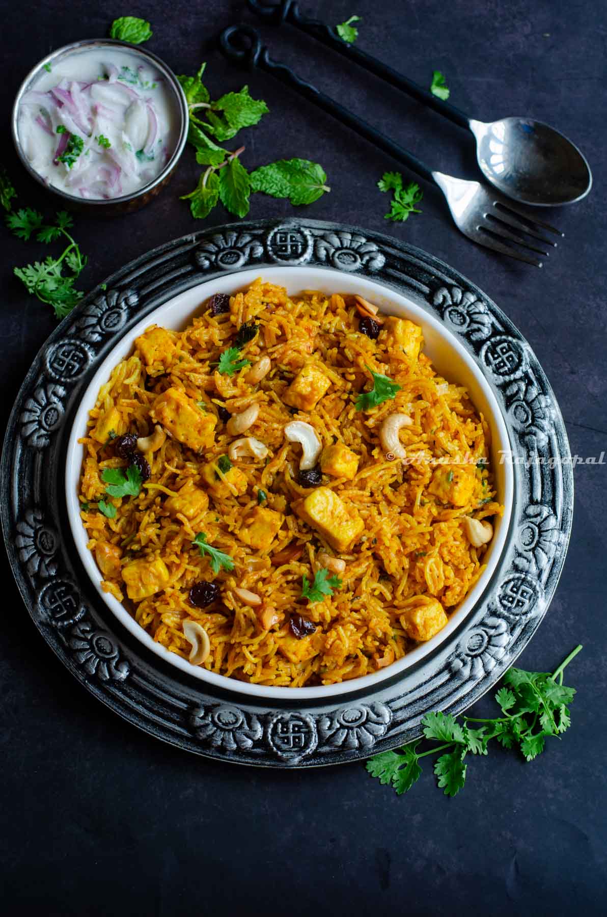 a-bowl-of-paneer-biryani-served-in-a-white-platter-with-onion-raita-and-cutlery-by-the-side